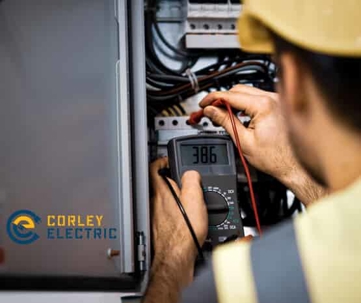 An electrician in a hard hat uses a multimeter to inspect an electrical panel.